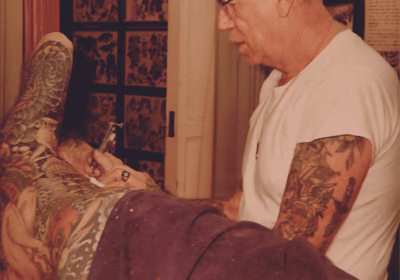 Vintage image of Sailor Jerry at work, inking a client in a traditional tattoo parlor setting.